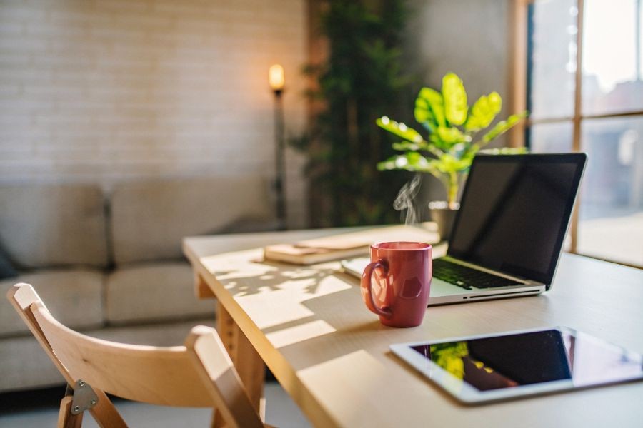 Shot of a beautiful home office set up with a chair and desk with a laptop open next to a red mug with a window and indoor plant in the distance.