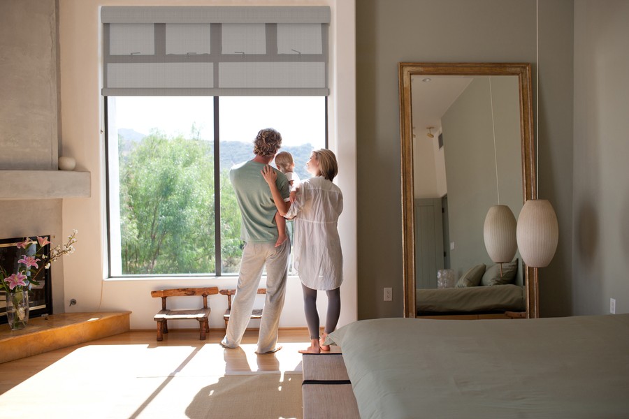 Family standing near a large window with motorized shades in a serene bedroom, featuring soft decor, a full-length mirror, and a cozy fireplace.