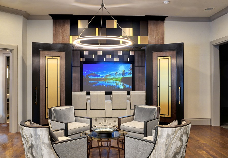 A luxurious home theater features tiered seating with plush chairs, a large wall-mounted screen, modern geometric wall paneling, and a stylish circular chandelier above a conversation area with elegant armchairs and a round coffee table.